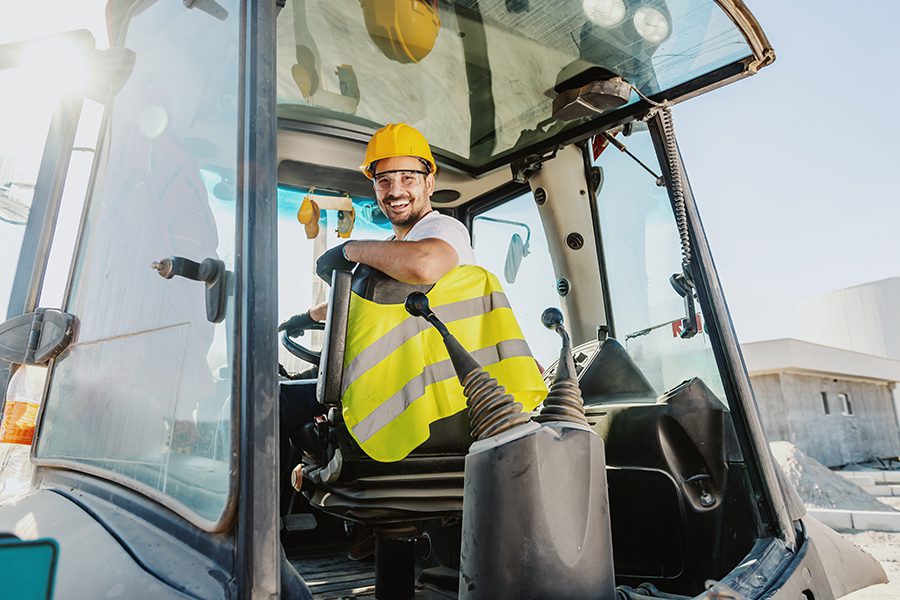 Specialized Business Insurance - Smiling Worker in Overalls with Helmet Driving an Excavator on Construction Site on a Bright Sunny Day